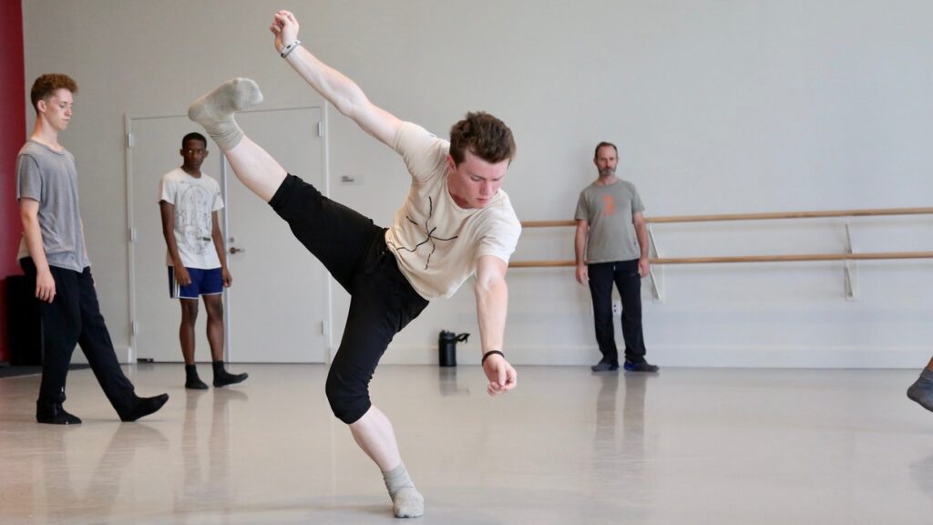 A man in a white shirt and black pants kicks his leg in the air in a dance studio.