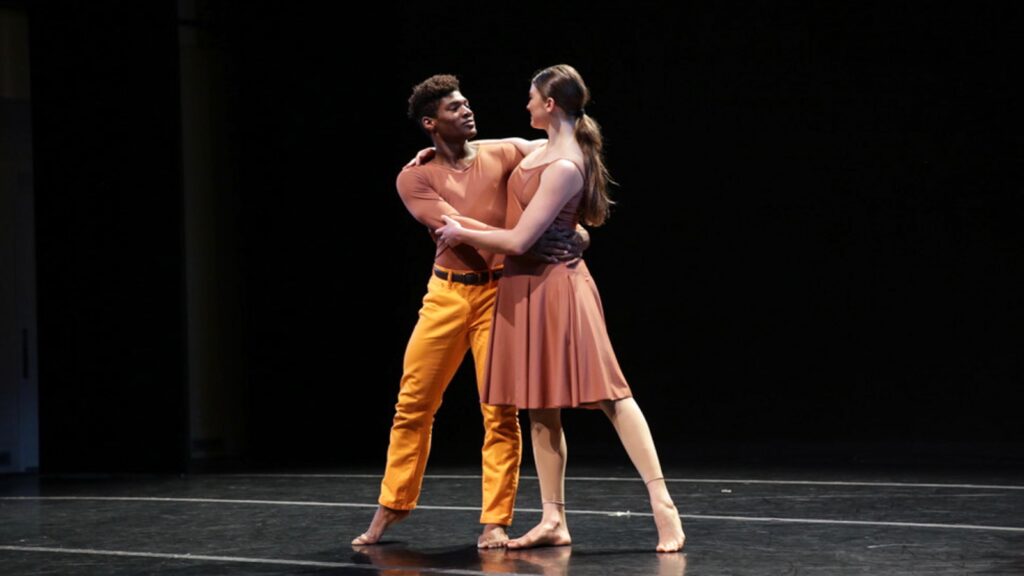 A man in a pale red shirt and orange pants embraces a woman on stage with a low ponytail wearing a pale red dress.