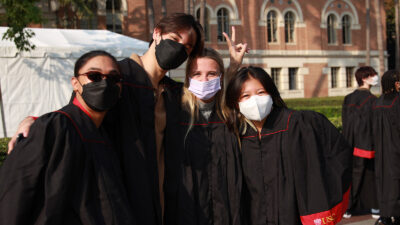 four students in black convocation robes pose for a picture. They are all wearing masks
