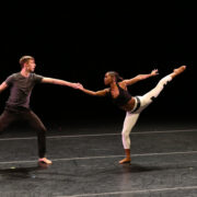 two dancers on stage, arms extended toward each other, dancer on the right with leg extended backwards