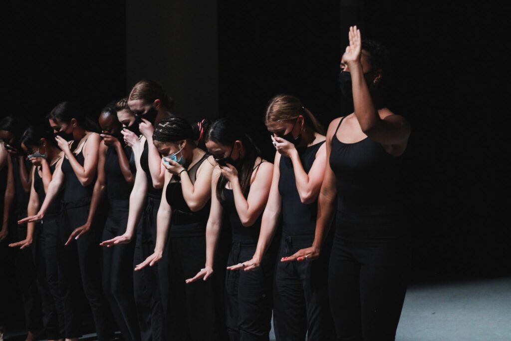 dancers lined up in a row, wearing all black, hands on their mouths