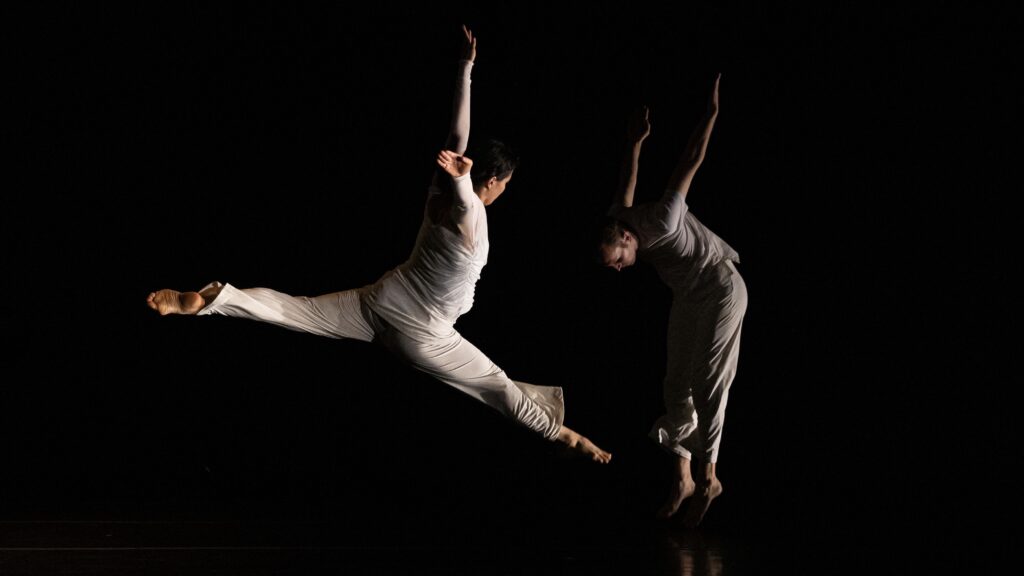 two dancers mid-jump against a black background, wearing long sleeves and pants