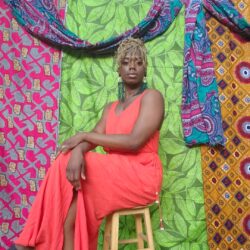 Toyin Sogunro sitting on a wooden stool in front of colorful tapestry wearing a coral dress