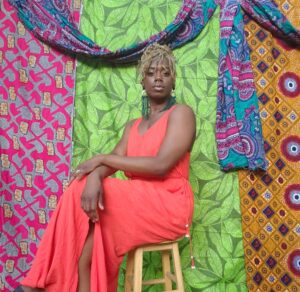 Toyin Sogunro sitting on a wooden stool in front of colorful tapestry wearing a coral dress