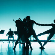 four dancers silhouetted in front of a group of musicians