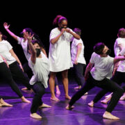 one dancer stands upright while a circle of dancer around her reach diagonally outwards. All are wearing white t-shirts and black pants