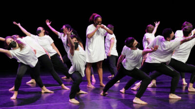 one dancer stands upright while a circle of dancer around her reach diagonally outwards. All are wearing white t-shirts and black pants