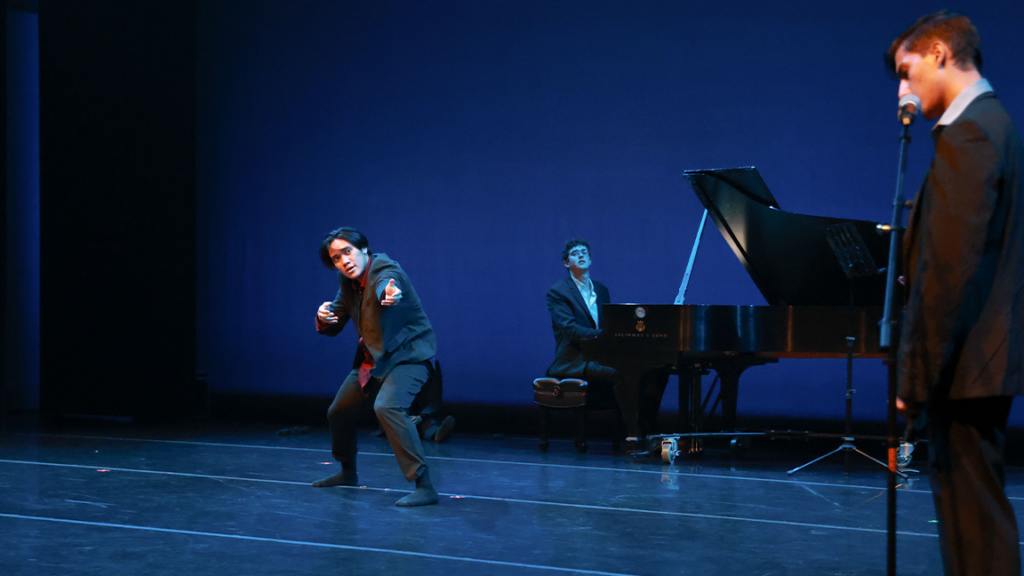 a dancer on stage. A man plays a grand piano in the back and another man speaks into a microphone on the right
