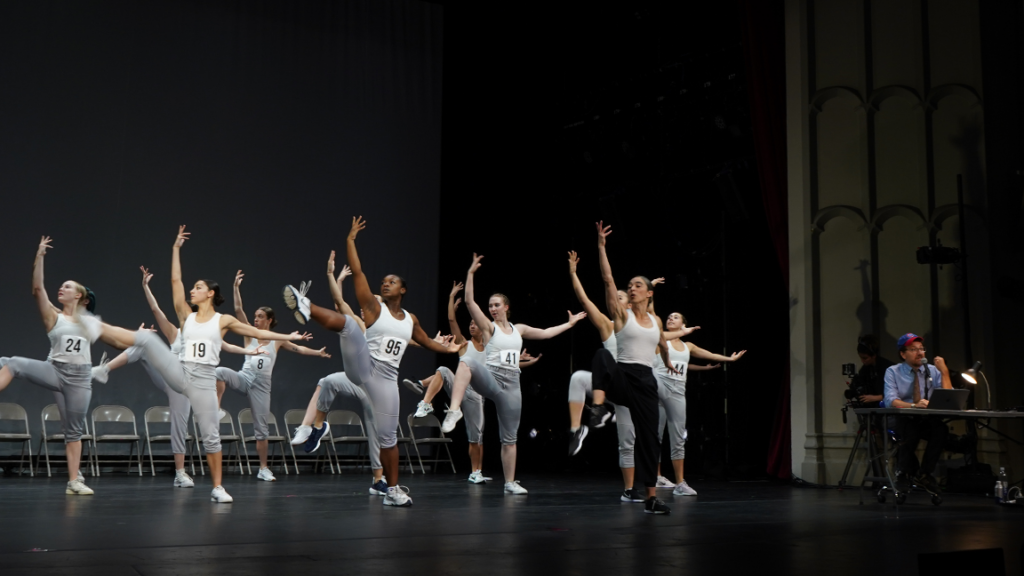 dancers on stage, all with one arm and one leg raised. Wearing white tops and gray pants with marathon numbers pinned to their shirts